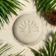 Round shampoo bar on sand surrounded by palm fronds