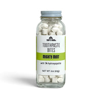 A clear glass jar of Soap A GoGo Toothpaste Bites in the Mighty Mint flavor, filled with white toothpaste bites and topped with a metal lid, set against a plain white background.