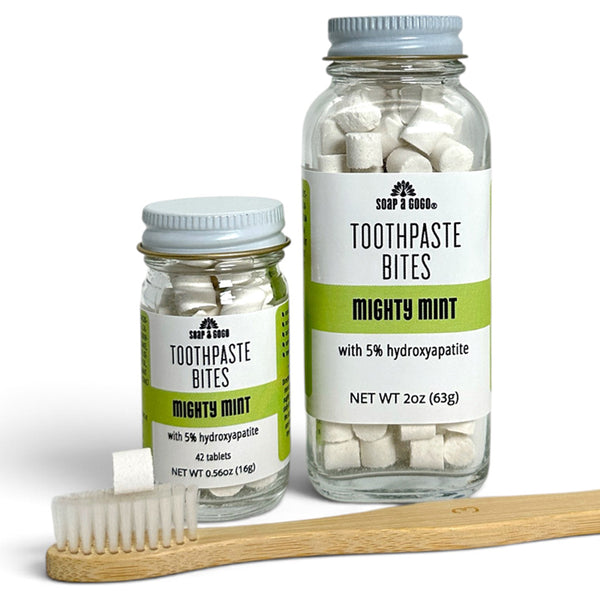 Two clear glass jars of Soap A GoGo Toothpaste Bites in Mighty Mint, one 2oz and one 0.56oz, with a wooden toothbrush resting in front on a white background.
