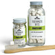Two clear glass jars of Soap A GoGo Toothpaste Bites in Mighty Mint, one 2oz and one 0.56oz, with a wooden toothbrush resting in front on a white background.